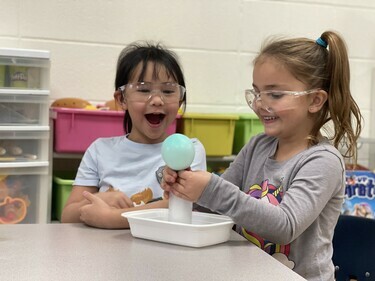 Two Kindergarten-aged girls with safety glasses on smiling playing with a ball on a cone in a bucket on a table